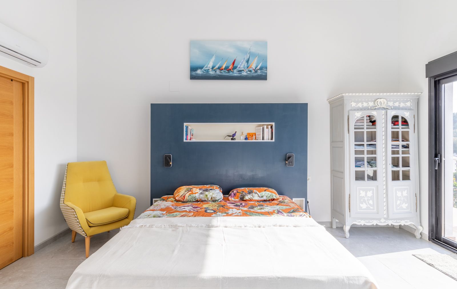 Modern bedroom featuring a blue integrated headboard with shelving, a yellow accent chair, and a vintage cabinet. Equipped with air conditioning and natural lighting.