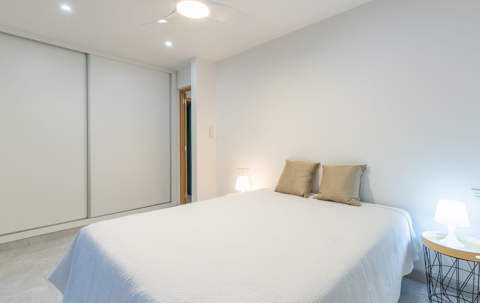 Modern bedroom featuring a large white sliding-door wardrobe, grey porcelain tile flooring, and integrated ceiling fan with recessed lighting.