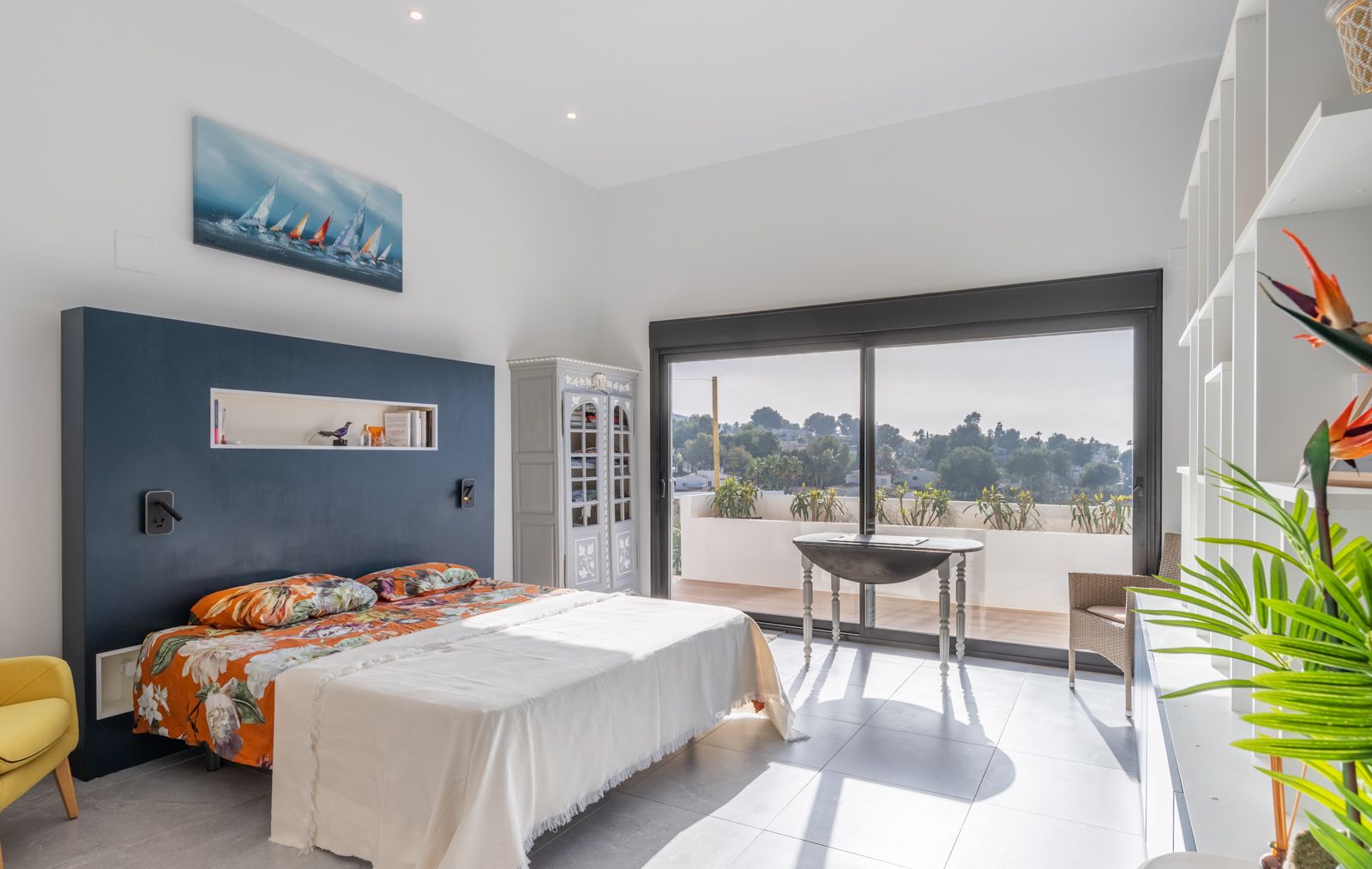 Modern bedroom featuring a navy blue accent wall with recessed shelving and large sliding doors opening onto a sunny terrace with open views.