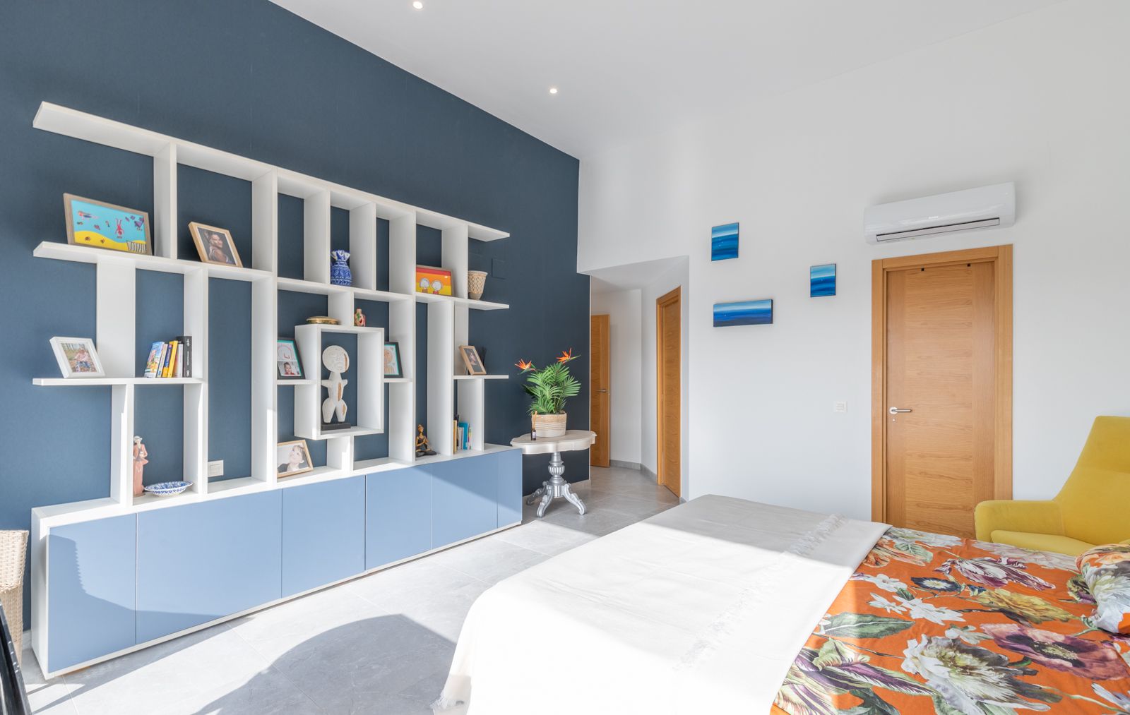 Modern bedroom featuring a white geometric shelving unit against a navy blue accent wall, integrated storage, and bright porcelain tile flooring.