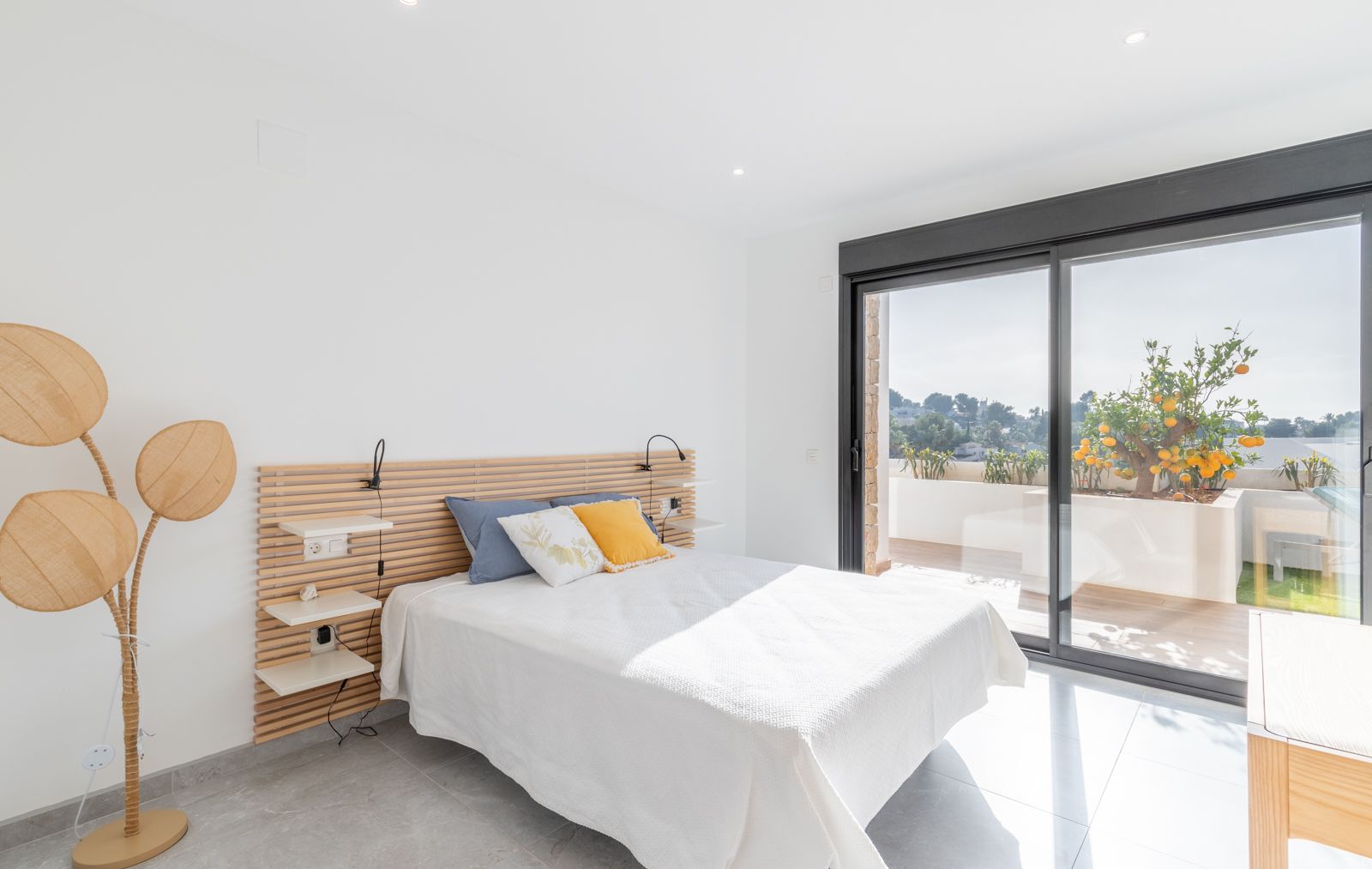 Modern bedroom featuring a wooden slatted headboard, integrated shelving, and large sliding doors opening onto a private terrace with citrus trees.