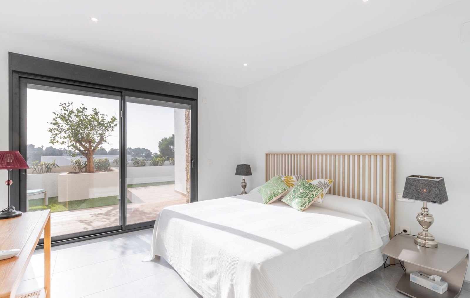 Modern bedroom featuring a wooden slatted headboard, large sliding glass doors opening to a private terrace, and recessed ceiling lighting.