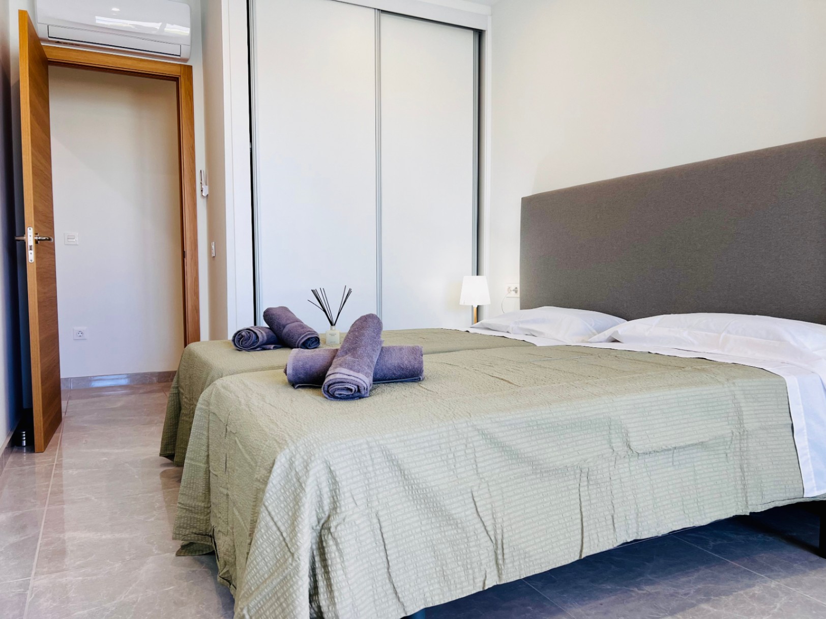 Modern bedroom featuring white built-in sliding wardrobes, porcelain tile flooring, and split-system air conditioning. Natural wood interior doors complement the neutral decor.