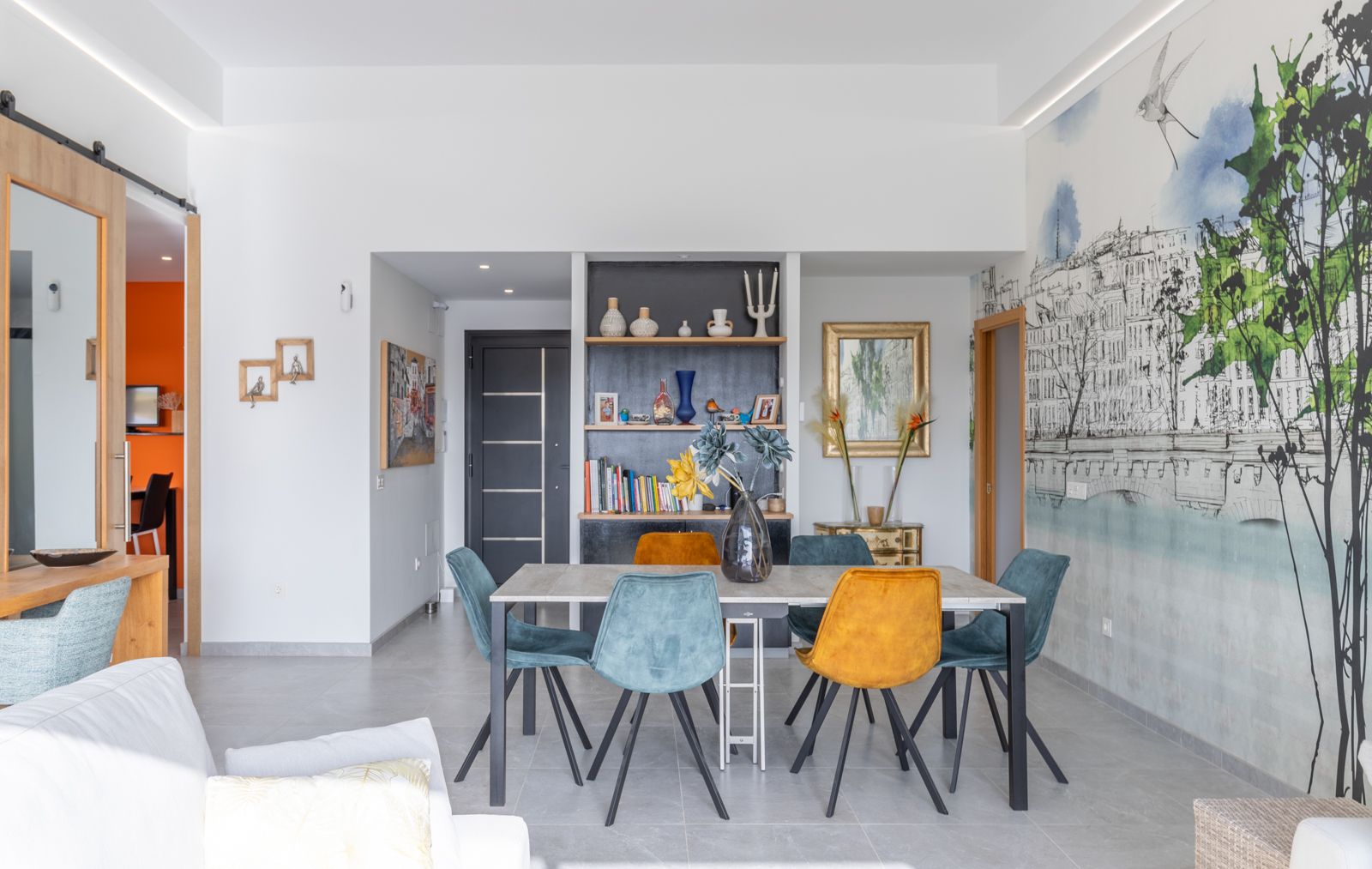 Modern dining area featuring a minimalist table with teal and mustard velvet chairs. The space includes a custom wall mural and built-in shelving under recessed LED lighting.