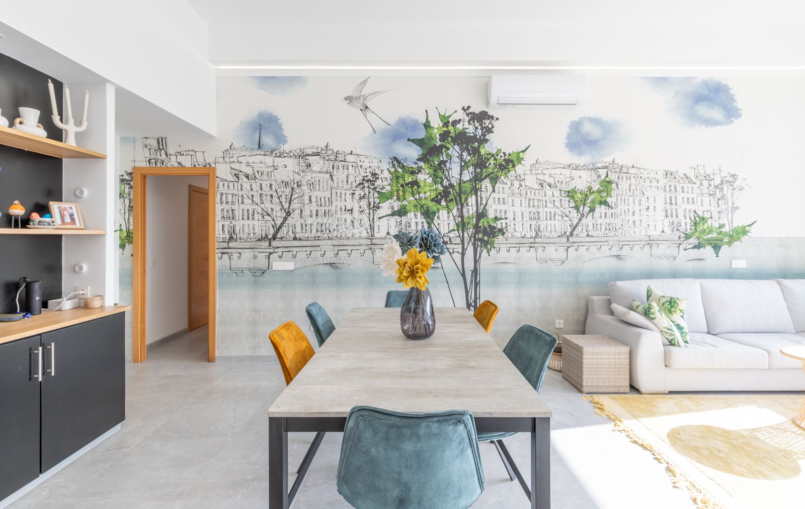Modern dining area featuring a stone-effect table, velvet chairs in teal and mustard, and a large urban mural. Grey tiled flooring and integrated living space.