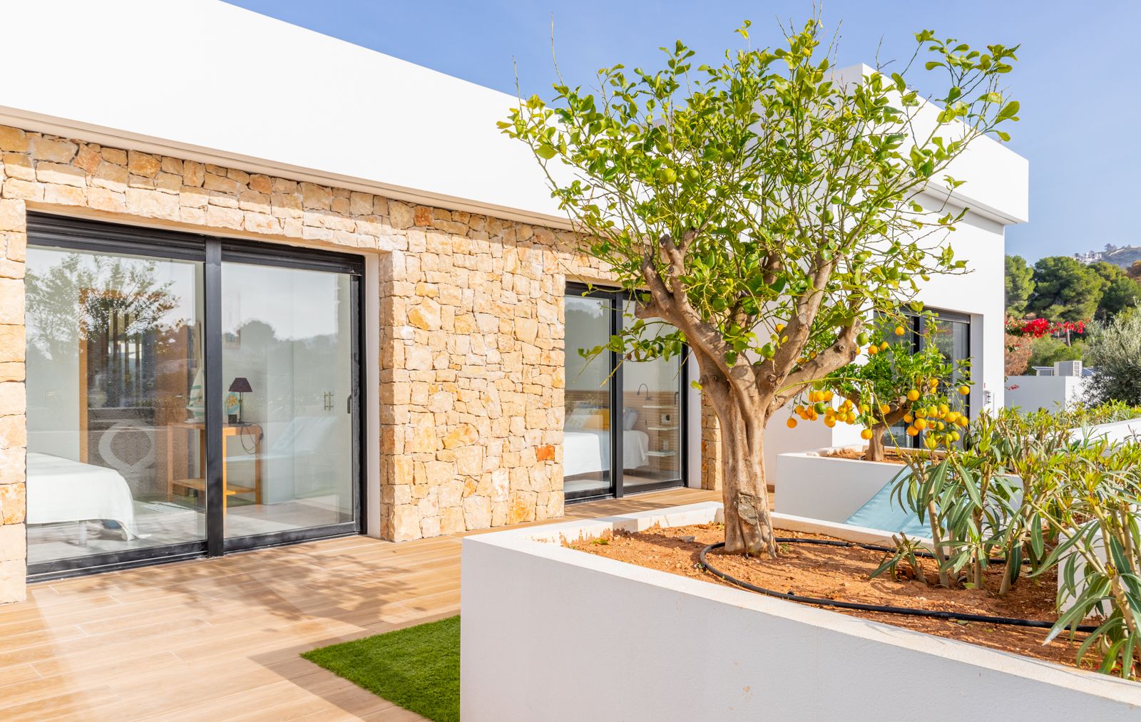 Modern facade featuring natural stone walls and floor-to-ceiling sliding glass doors that open onto a wood-effect terrace with integrated Mediterranean planters.