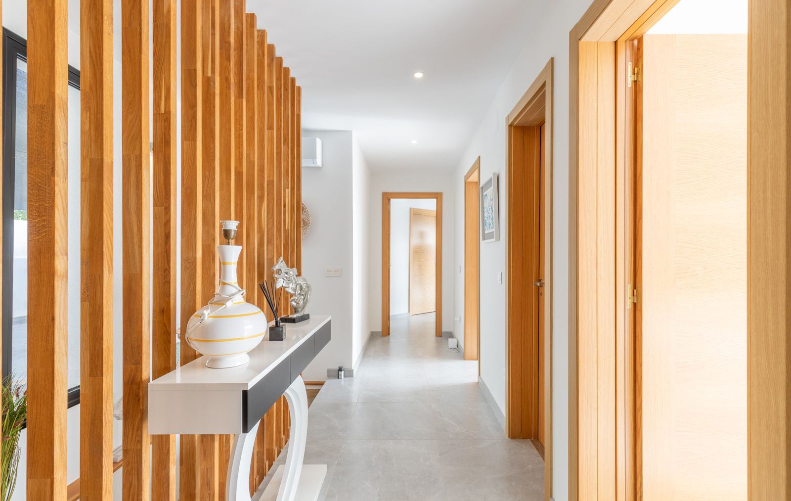 Modern hallway featuring a vertical wooden slat room divider, grey porcelain flooring, and matching light wood door frames with recessed lighting.
