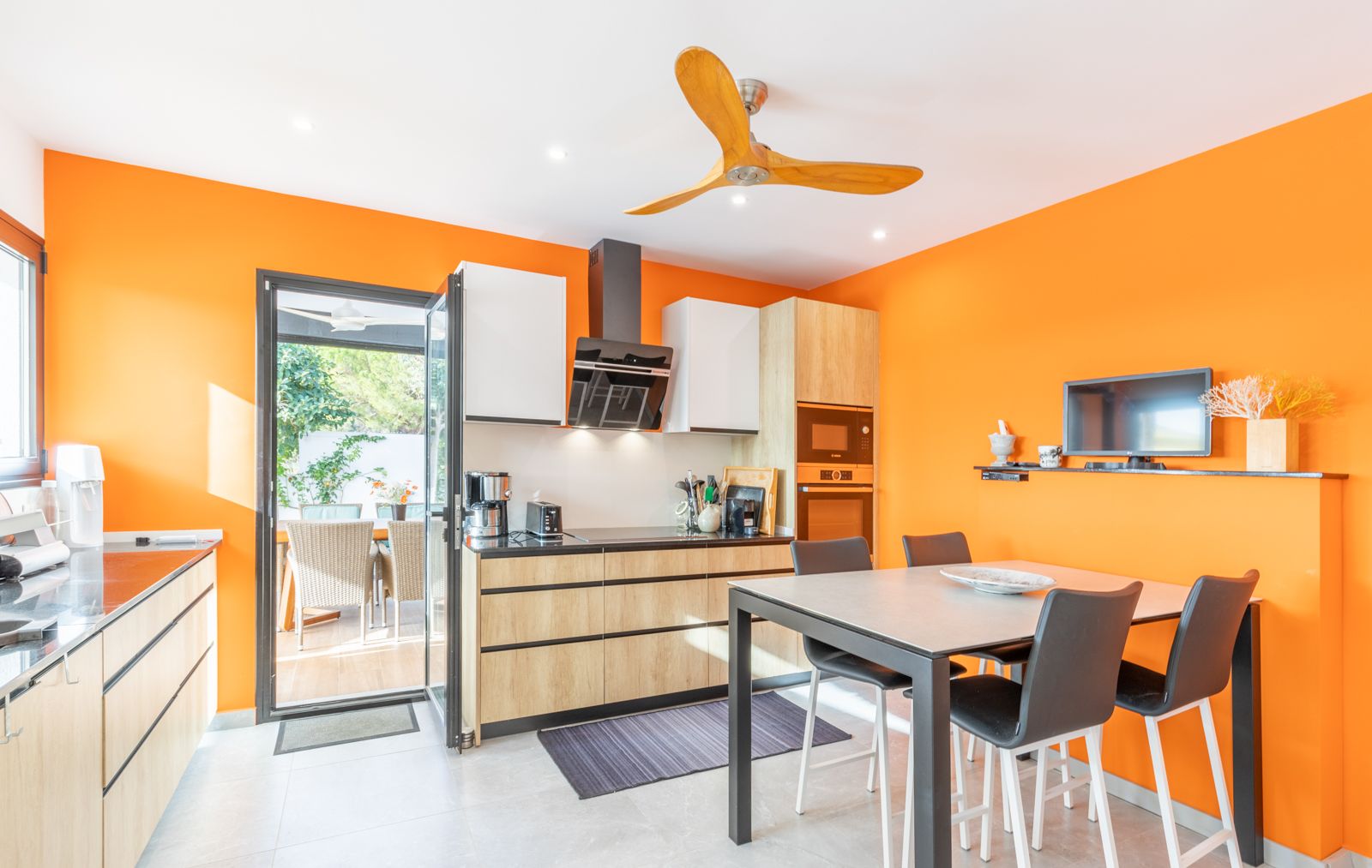 Modern kitchen featuring vibrant orange walls, light wood cabinetry, and an integrated dining area with direct access to the outdoor terrace.