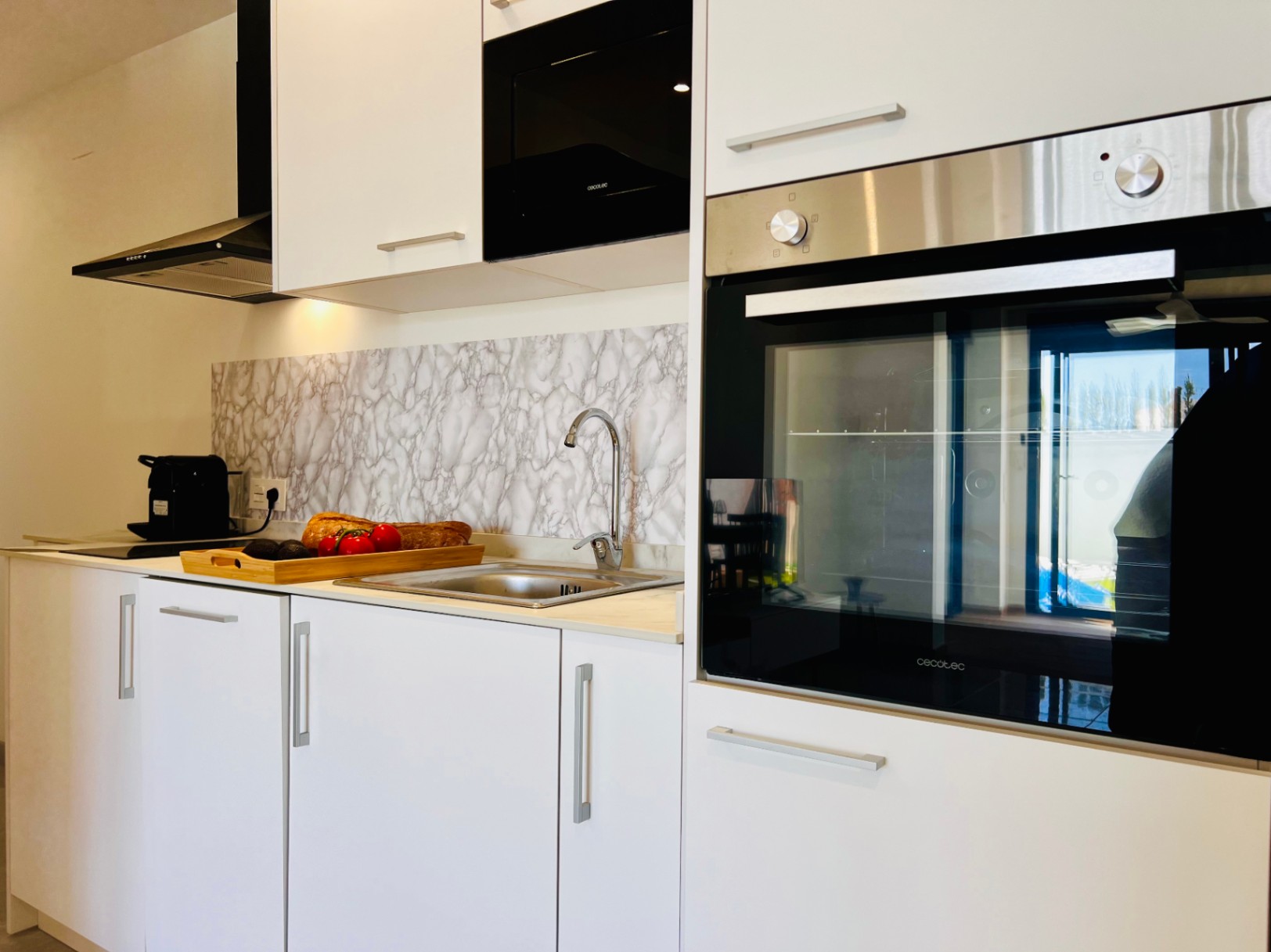 Modern kitchen featuring white cabinetry, integrated Cecotec appliances, and a marble-effect backsplash. It includes a stainless steel sink and a sleek black extractor hood.