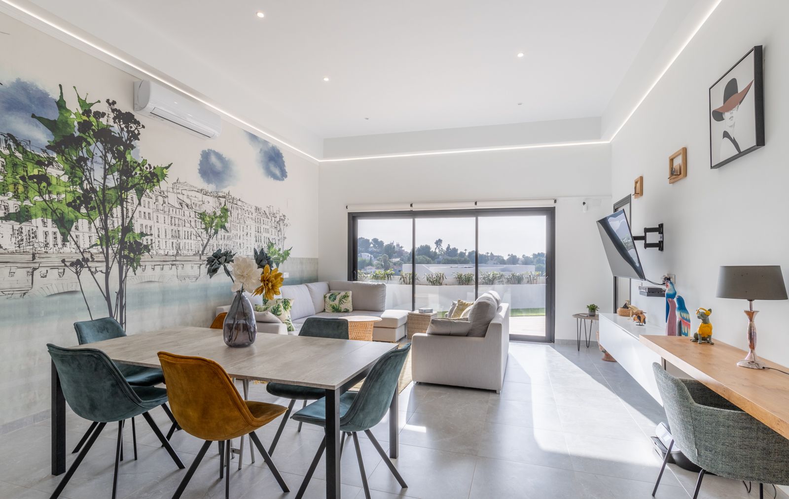Modern living and dining area featuring a custom wall mural, contemporary velvet seating, and large sliding doors opening to a sunlit terrace.