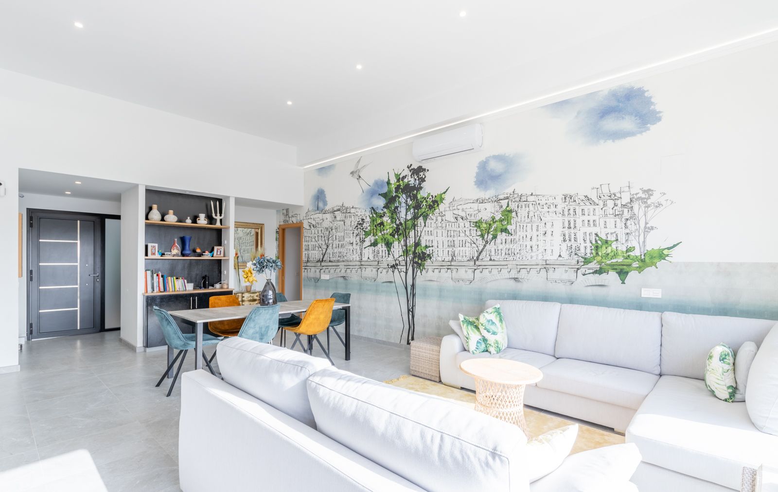 Open-plan living and dining area featuring a large sectional sofa, a decorative urban mural, and contemporary furniture under recessed ceiling lighting.