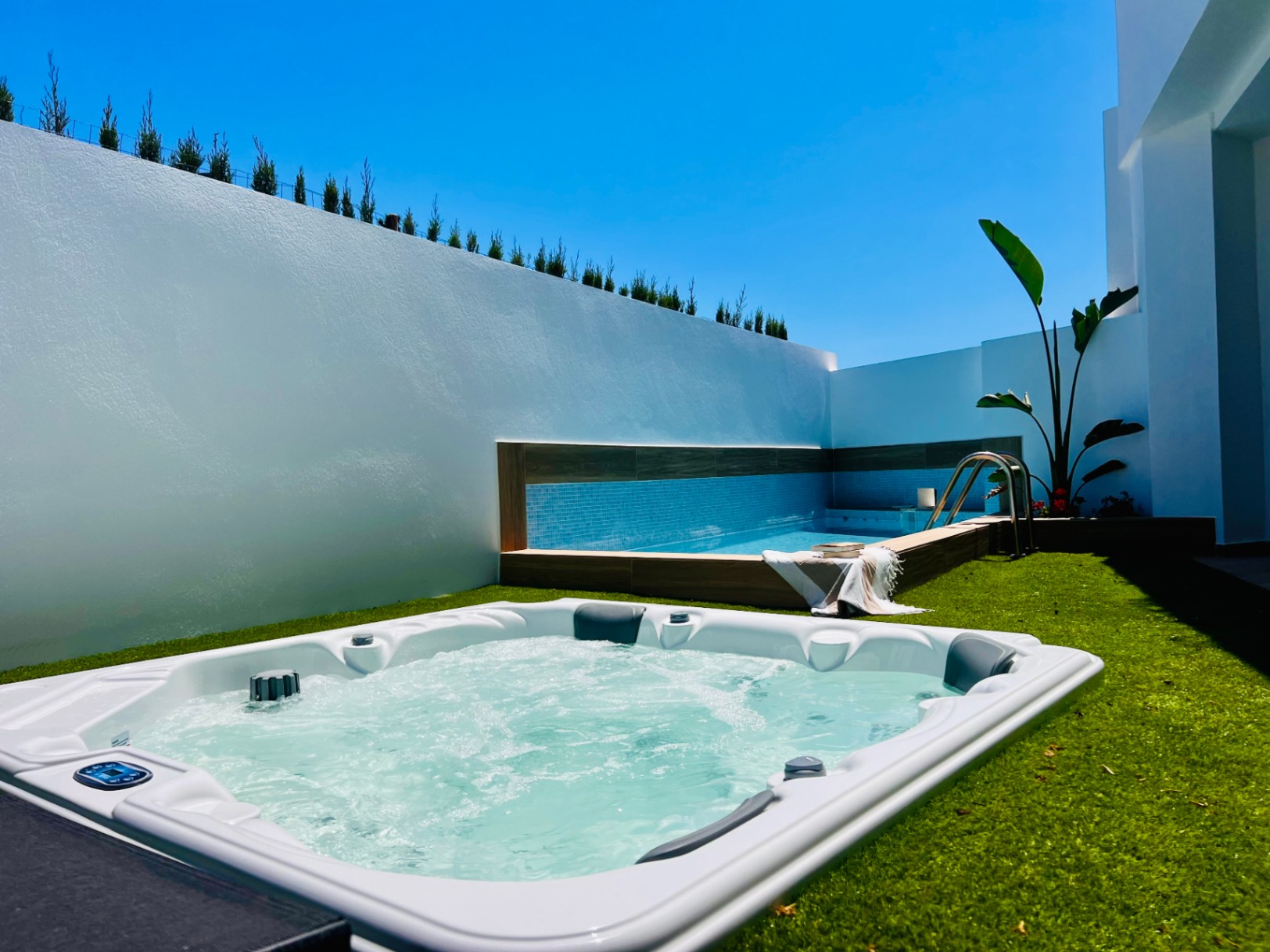 Outdoor hot tub embedded in artificial turf, located next to a private pool with blue tiling and high white walls for complete privacy.