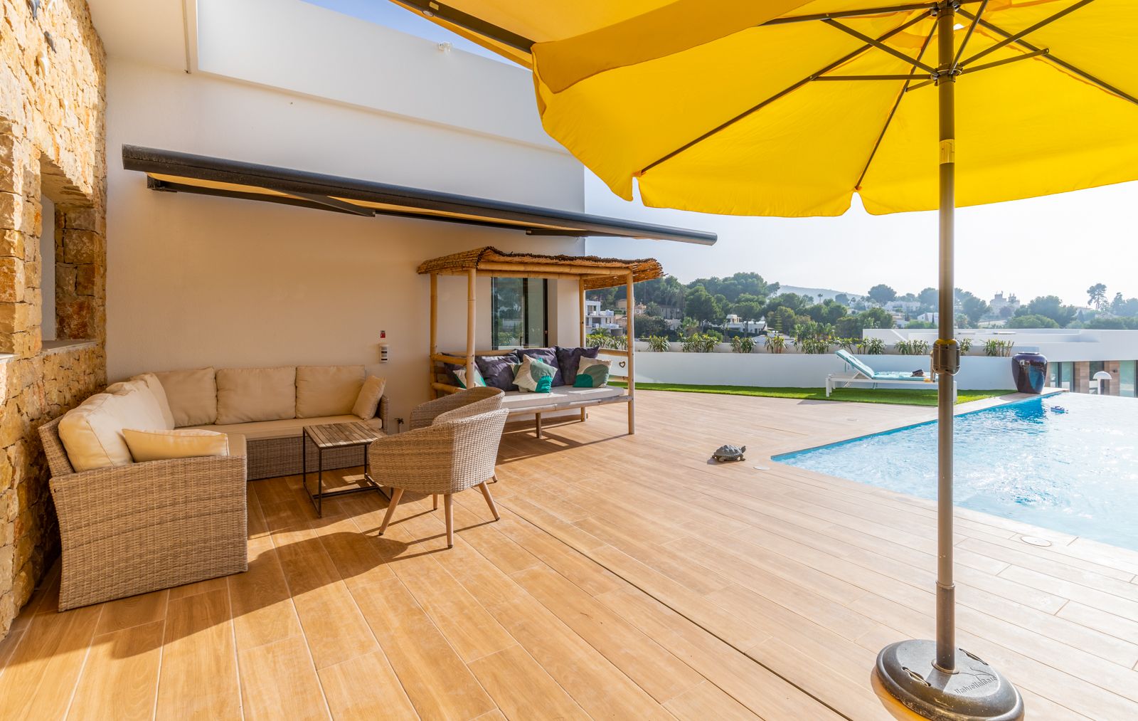 Outdoor terrace featuring wood-effect porcelain flooring, wicker lounge seating, a bamboo daybed, and a swimming pool framed by natural stone walls and a yellow parasol.
