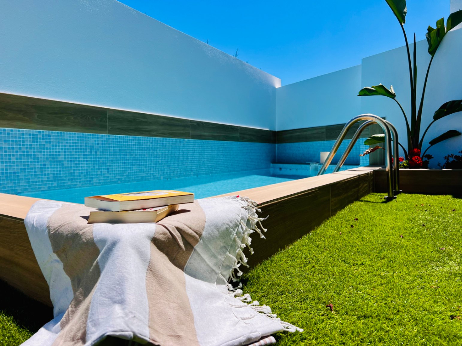Private jacuzzi featuring blue mosaic tiling, wood-effect trim, and an adjacent artificial lawn area under a clear Mediterranean sky.