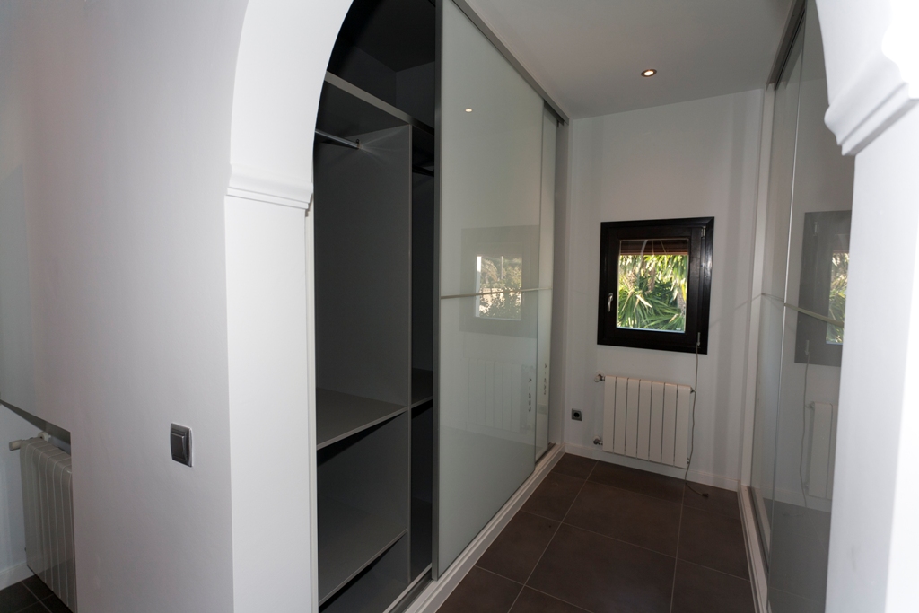 Dressing area featuring built-in wardrobes with frosted glass sliding doors, interior shelving, dark tiled flooring, radiator heating, and recessed ceiling lights.