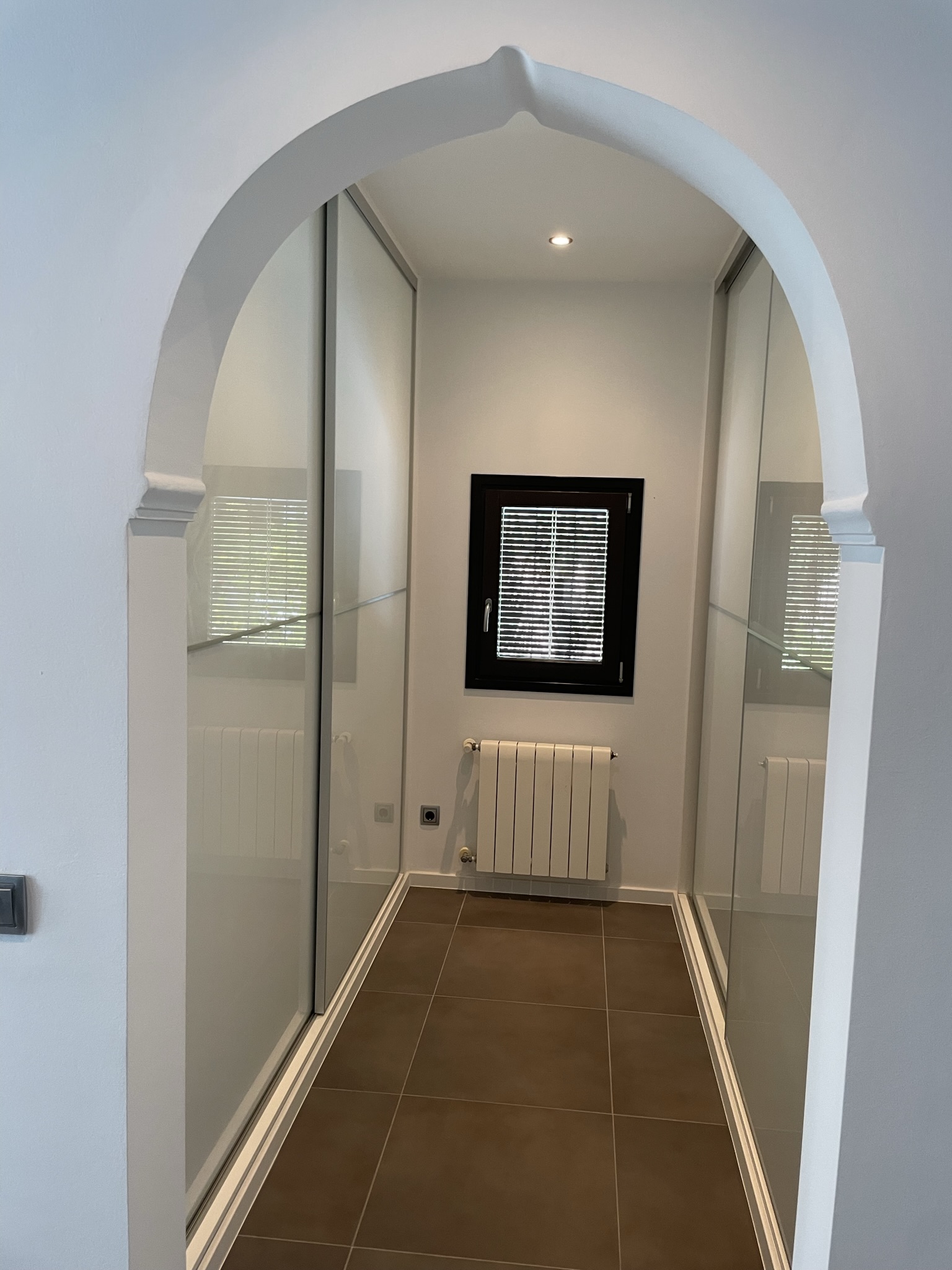 Dressing area featuring built-in wardrobes with sliding doors, accessed through a traditional pointed archway and equipped with a central heating radiator.