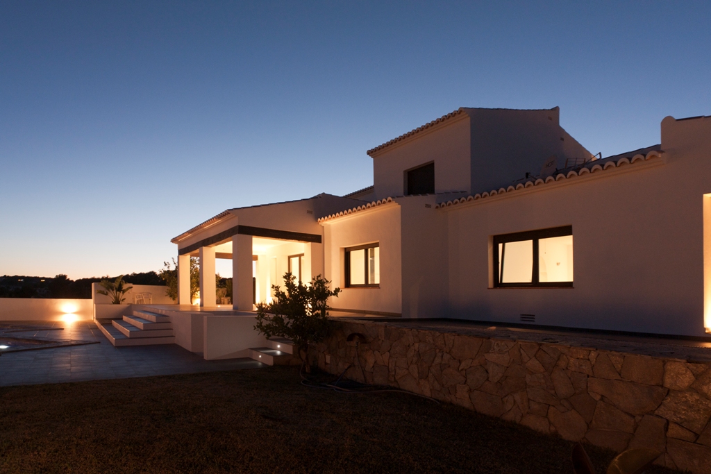 Exterior facade of the villa at twilight, featuring warm ambient lighting, natural stone walls, and spacious terraces leading to the pool area.