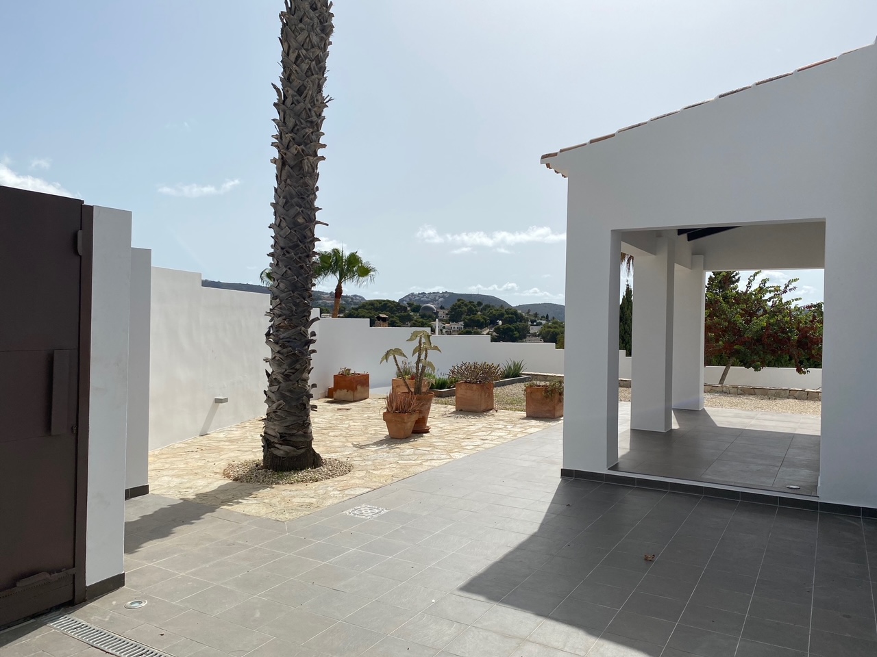 Exterior patio featuring grey ceramic tiles and natural stone paving, accented by a tall palm tree and terracotta planters with views of the surrounding hills.