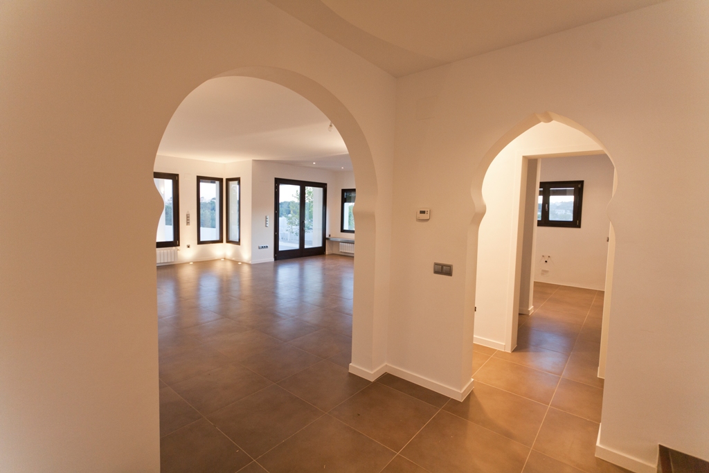 Hallway featuring Mediterranean-style arches, terracotta-toned floor tiles, and a view into the living area with dark-framed aluminum windows.