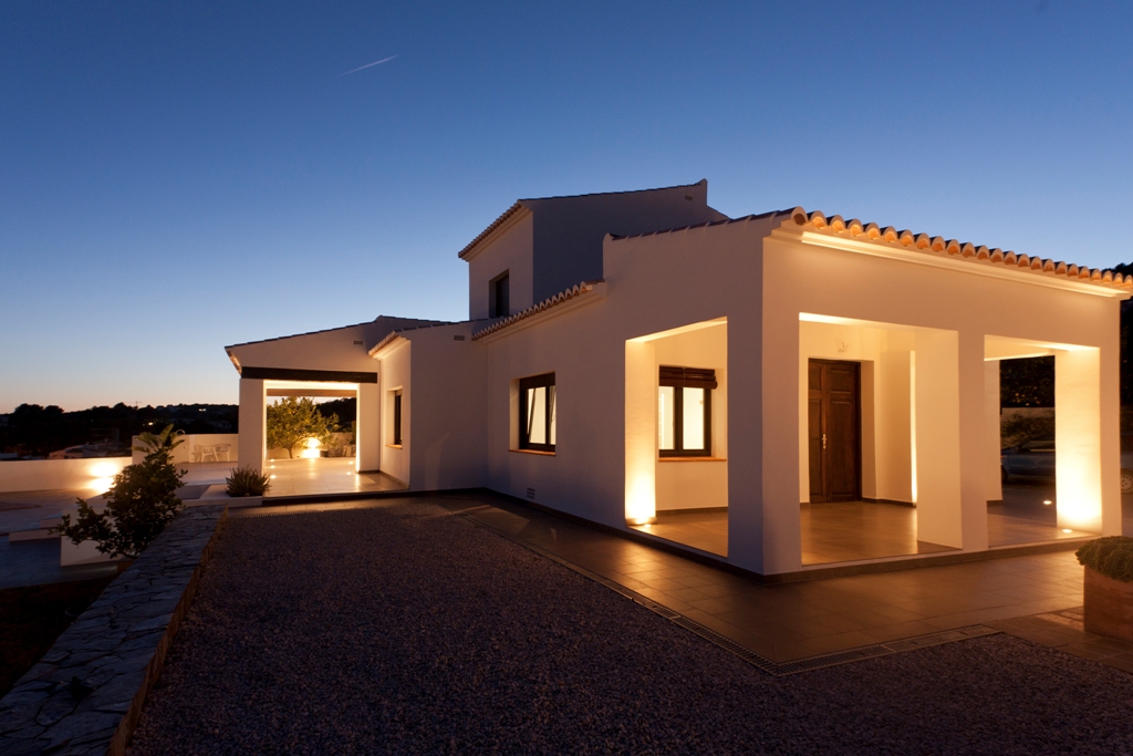 Mediterranean facade at dusk featuring integrated floor lighting on pillars, highlighting the restored wooden entrance door and white exterior walls.