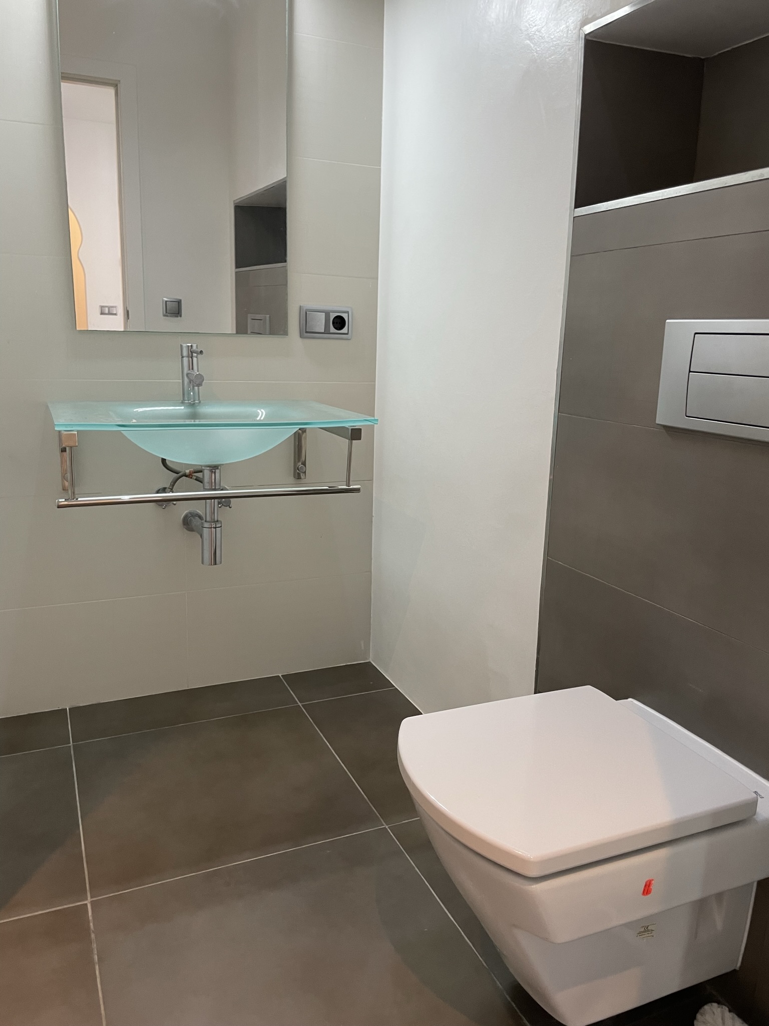 Modern bathroom featuring a translucent glass washbasin with chrome supports and a wall-hung toilet. Large-format grey and cream tiles create a clean aesthetic.