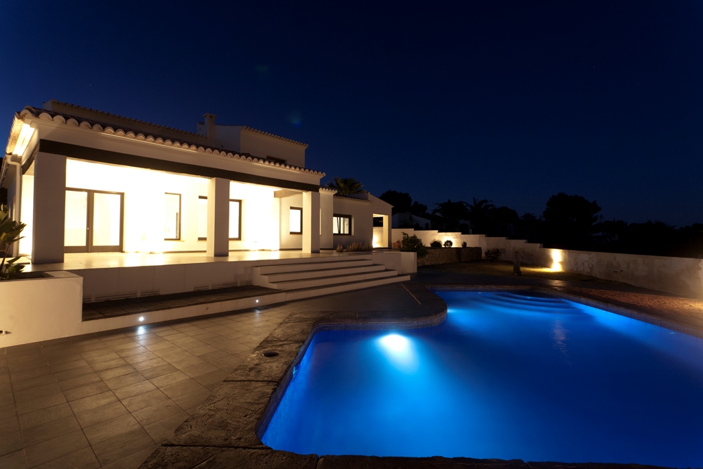 Night view of the illuminated white facade and the irregular-shaped pool with blue underwater lighting, set against a spacious tiled terrace.
