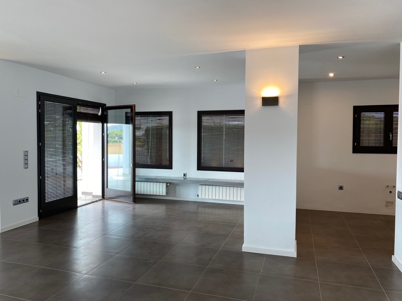 Open-plan living area featuring dark tiled flooring, recessed ceiling lights, and wall-mounted radiators. Large windows with integrated blinds provide ample natural light.