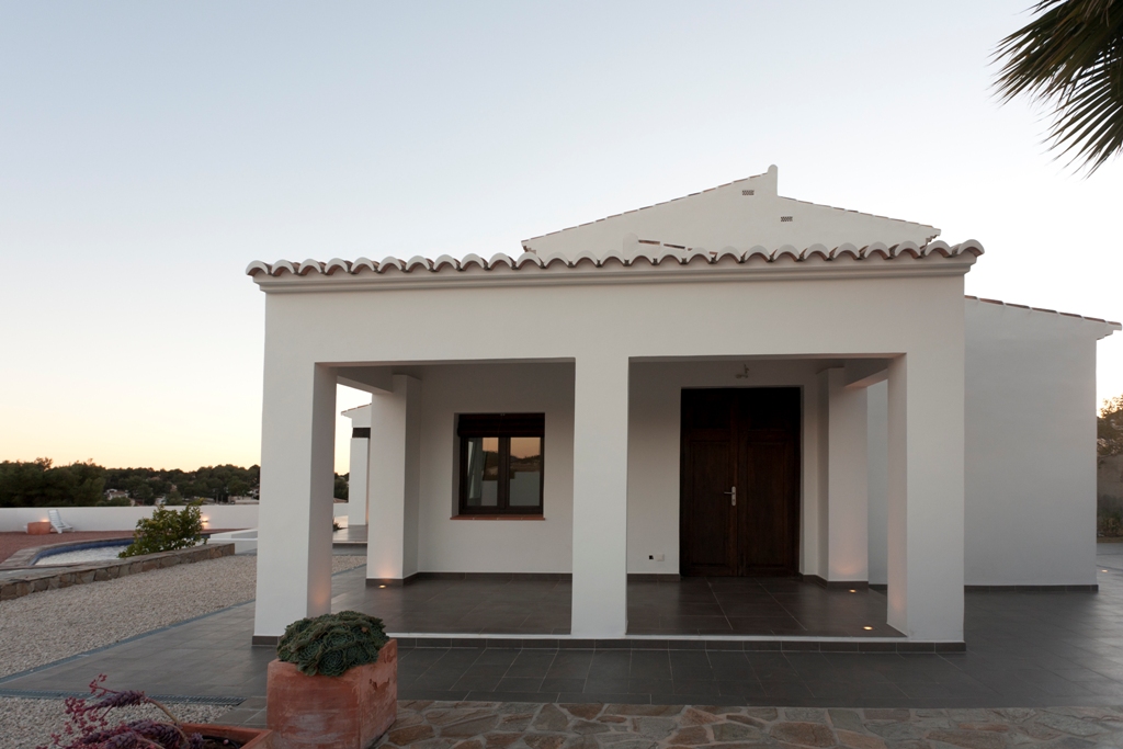 White facade featuring a covered porch with integrated floor lighting and a restored antique wooden door, set beside a gravel area and pool.
