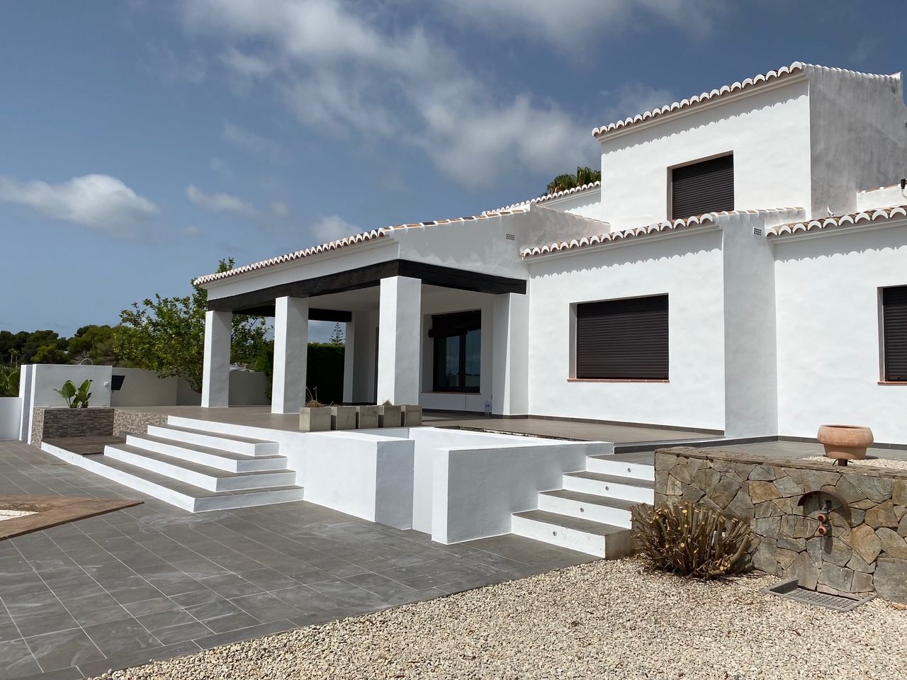 White Mediterranean facade featuring dark wooden beams, a spacious tiled terrace, and natural stone walls. Includes an outdoor shower area and integrated access steps.