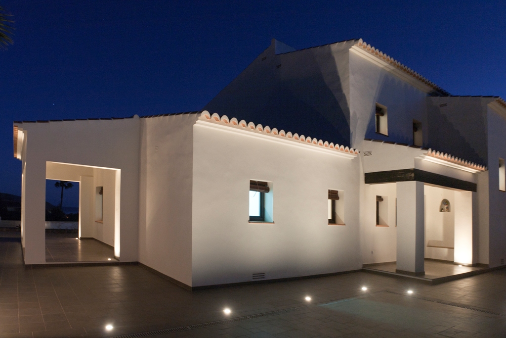 White Mediterranean facade illuminated at dusk with recessed floor spotlights. Features include traditional terracotta roof tiles and a covered porch with dark wooden beams.