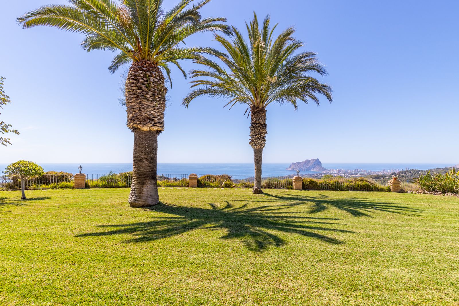 View of the garden with green lawn, palm trees and views of the Mediterranean Sea and the Peñón de Ifach.