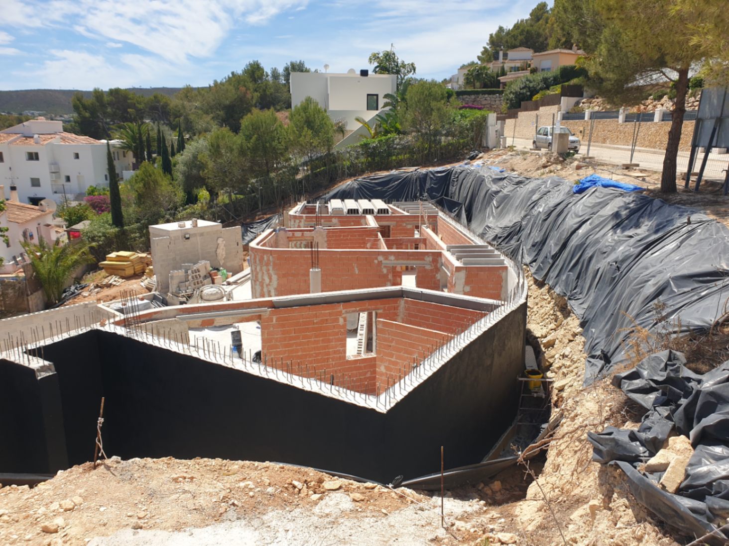 Construction progress showing the brick structure and waterproofed retaining walls on a sloped plot within a residential area of Jávea.