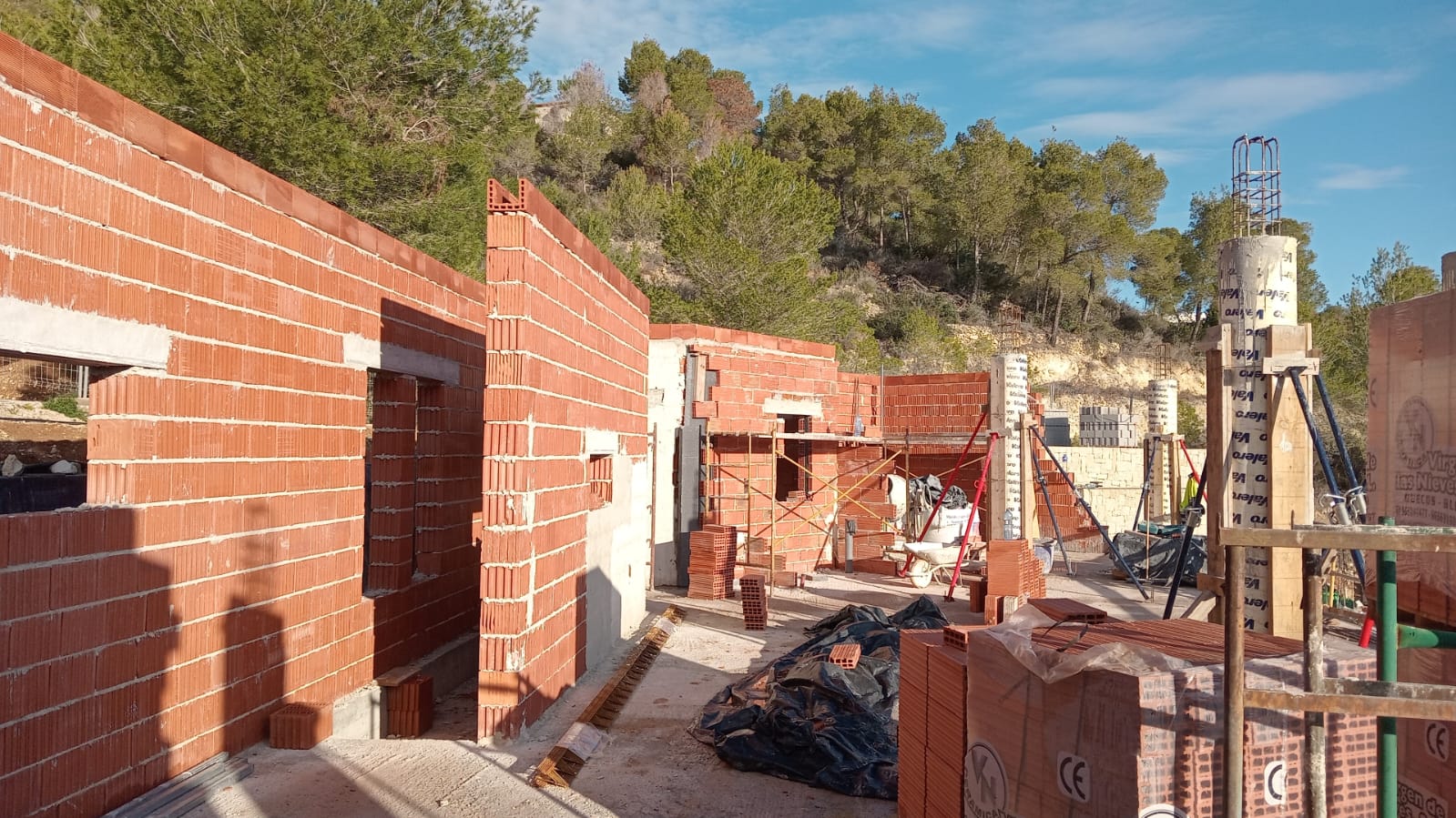 Current construction phase featuring termoarcilla brick walls and concrete pillars set against a backdrop of Mediterranean pine forest.