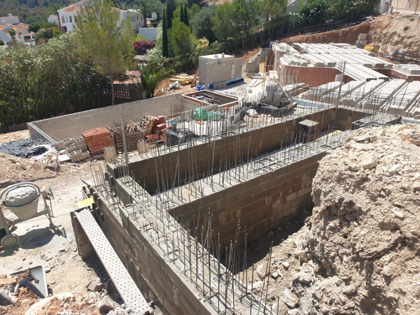Early construction phase showing foundation walls with steel reinforcement and concrete blocks on an elevated plot surrounded by Mediterranean greenery.