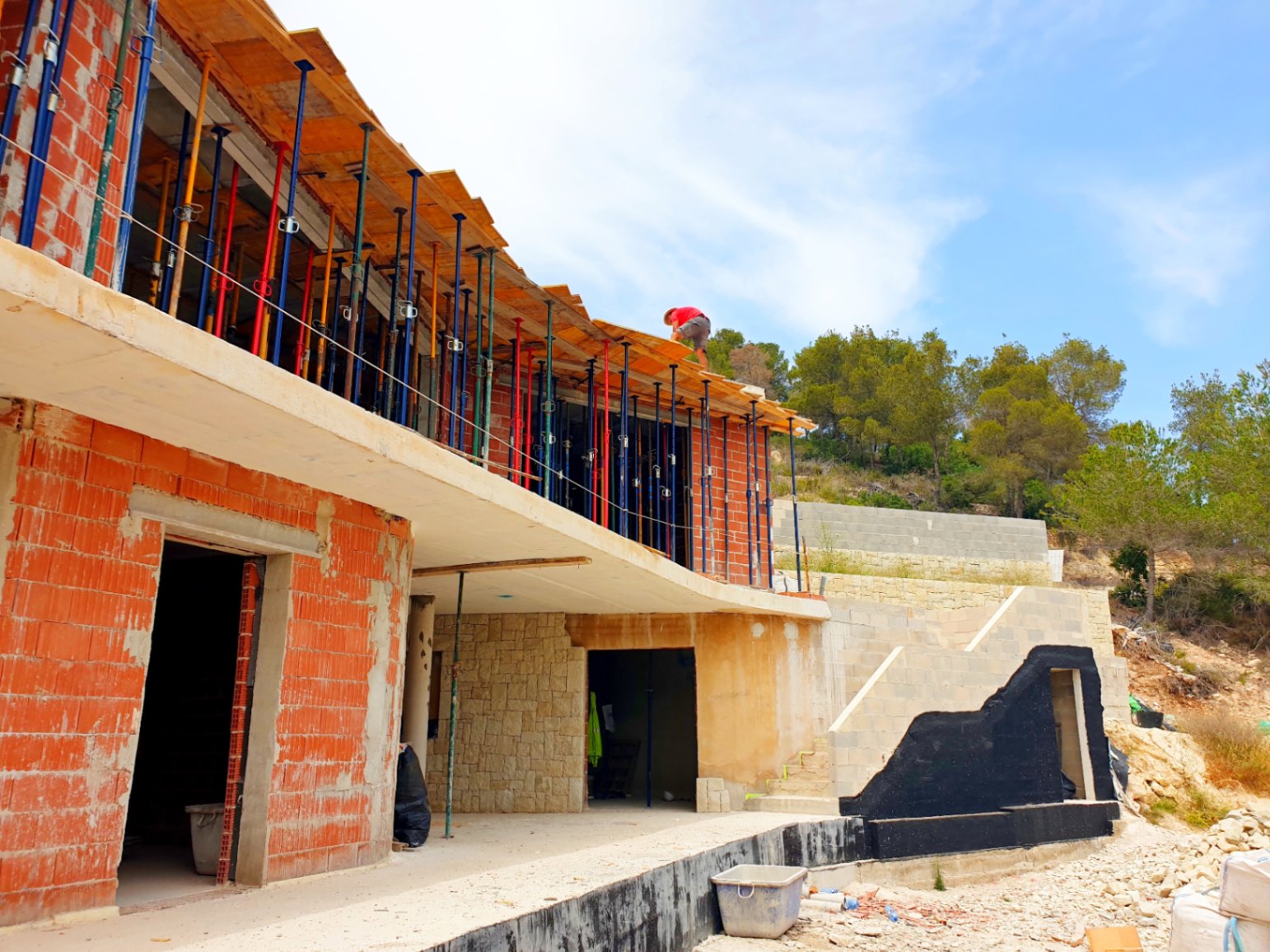 Exterior view of the villa under construction, featuring the brick structure, concrete slabs with shoring props, and integrated natural stone wall sections.