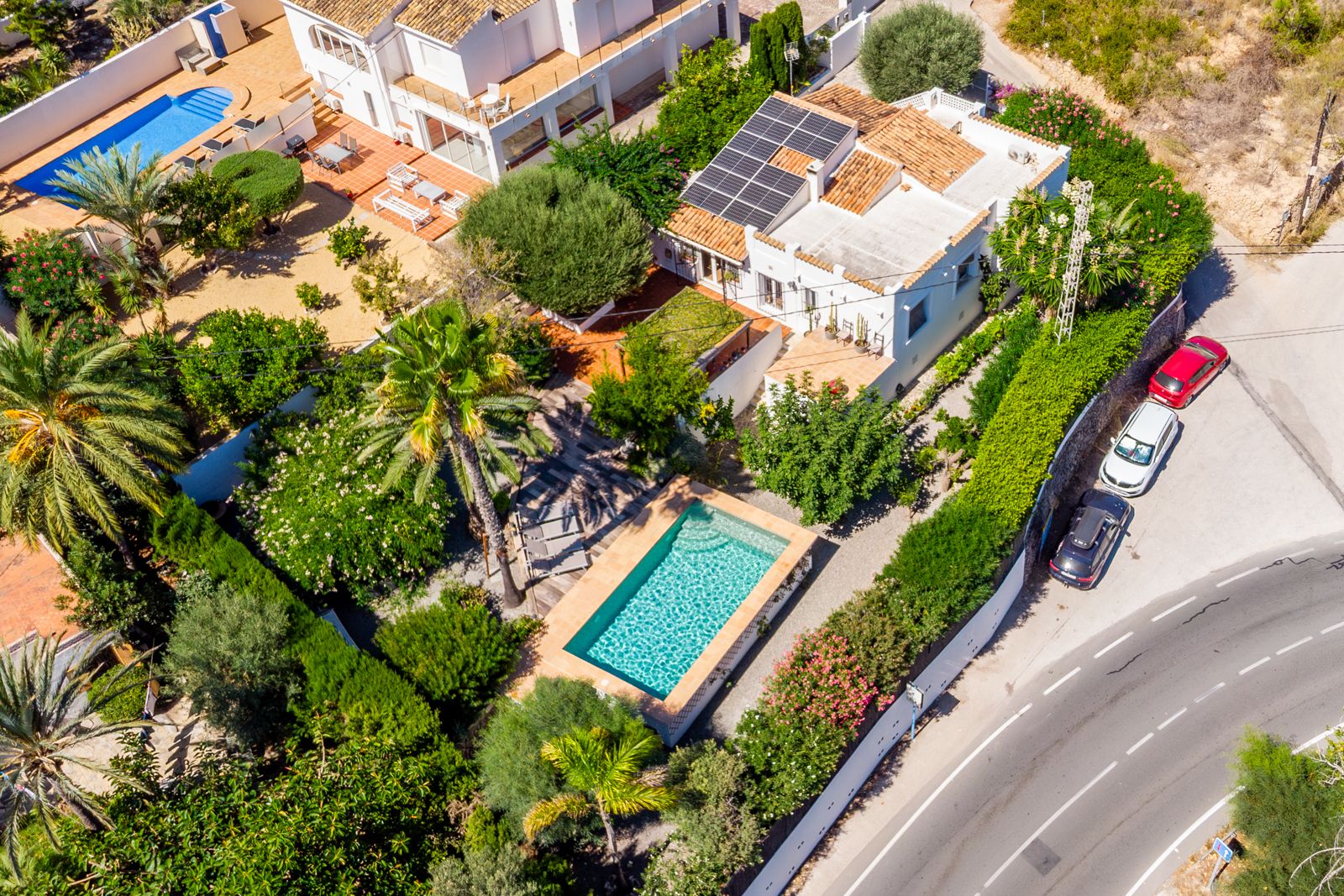 Aerial perspective of the villa showcasing the rectangular swimming pool, lush mature gardens, and the solar panel system on the terracotta roof.