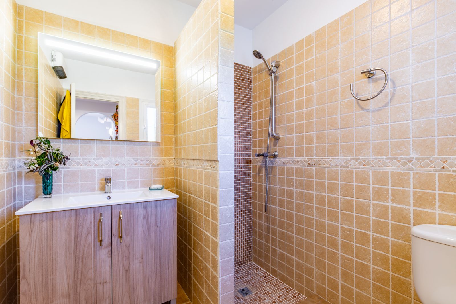 Bathroom featuring a walk-in shower with mosaic flooring and warm beige tiling. A wood-effect vanity unit and backlit mirror complete the functional space.
