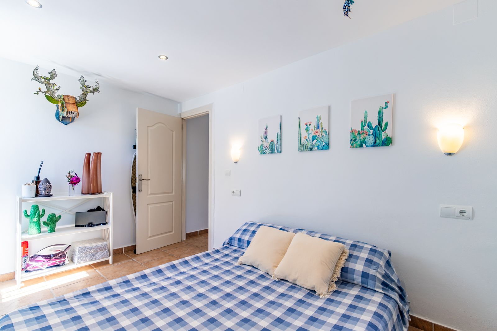 Bright bedroom featuring terracotta tile flooring, a bed with blue checkered linens, and cactus-themed decor. Equipped with recessed lighting and warm wall sconces.