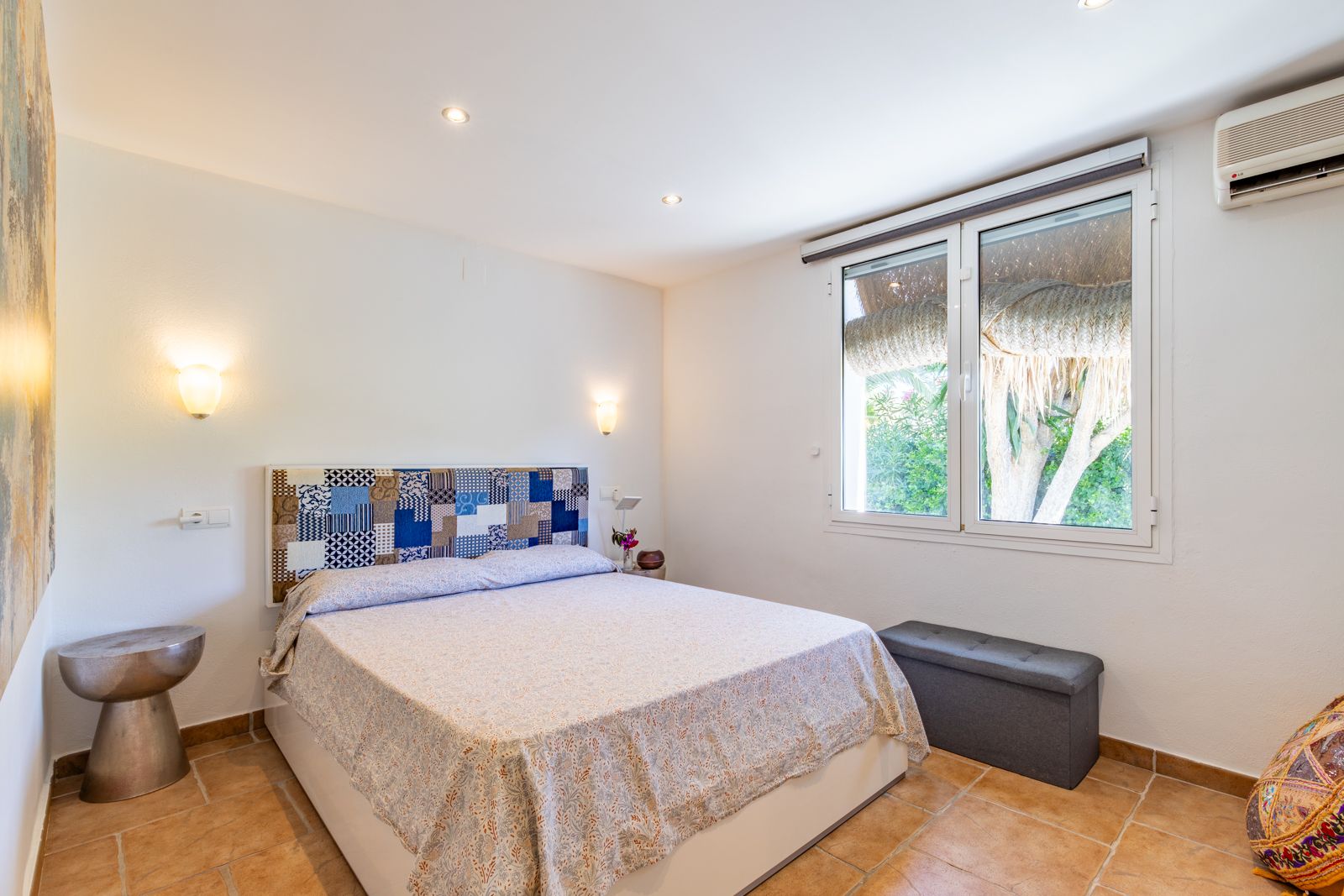 Bright bedroom featuring terracotta tile flooring, a patchwork headboard, air conditioning, and a window with views of the garden.
