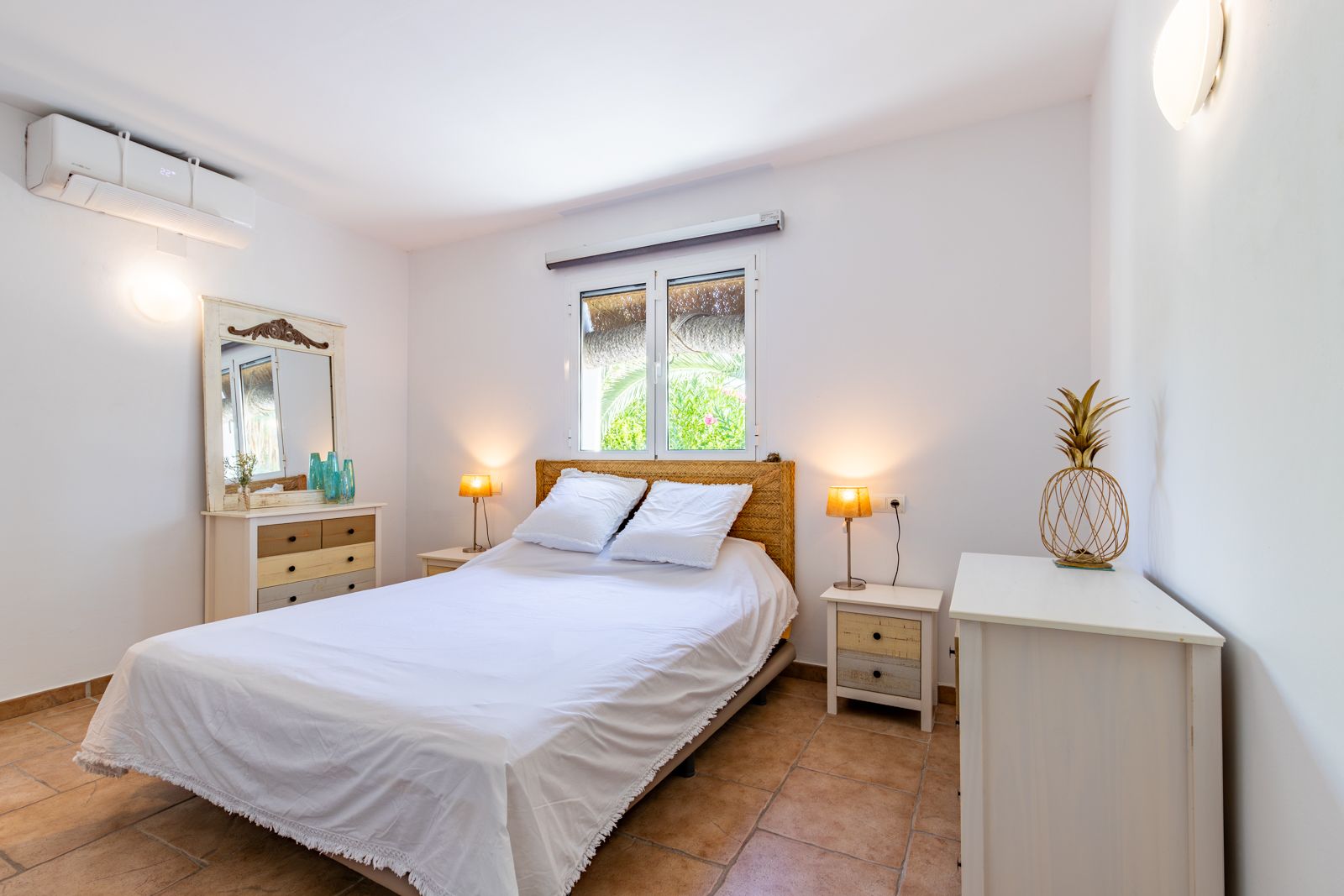 Bright bedroom featuring terracotta tile flooring, a split air conditioning unit, and rustic wooden furniture. The space includes a natural fiber headboard and white-framed windows.