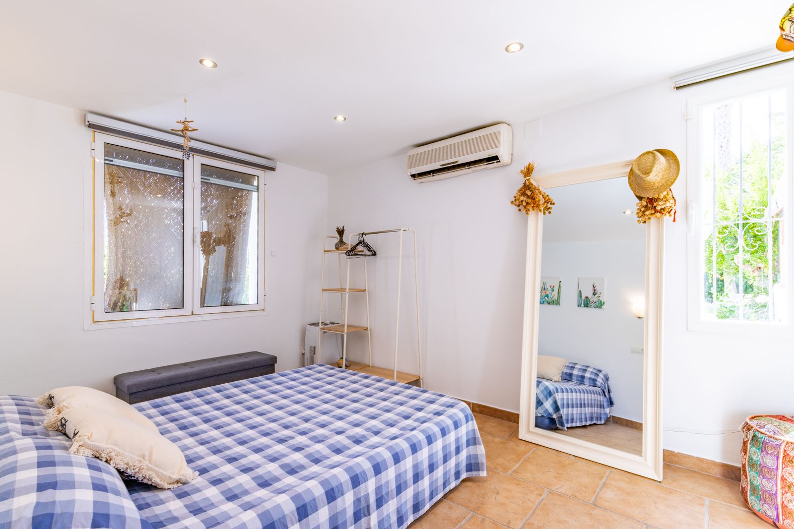 Bright bedroom featuring terracotta tile flooring, split air conditioning, and a large floor mirror. Recessed ceiling lights and natural light through barred windows.