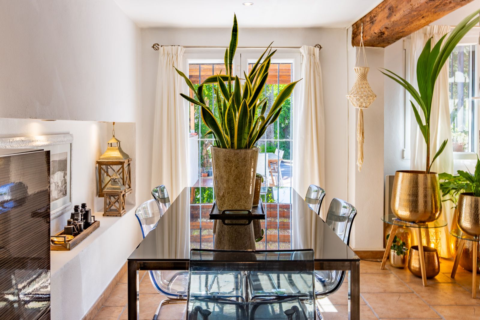Bright dining area featuring a reflective glass table, transparent chairs, and exposed wooden beams. Large French doors provide direct access to the garden.