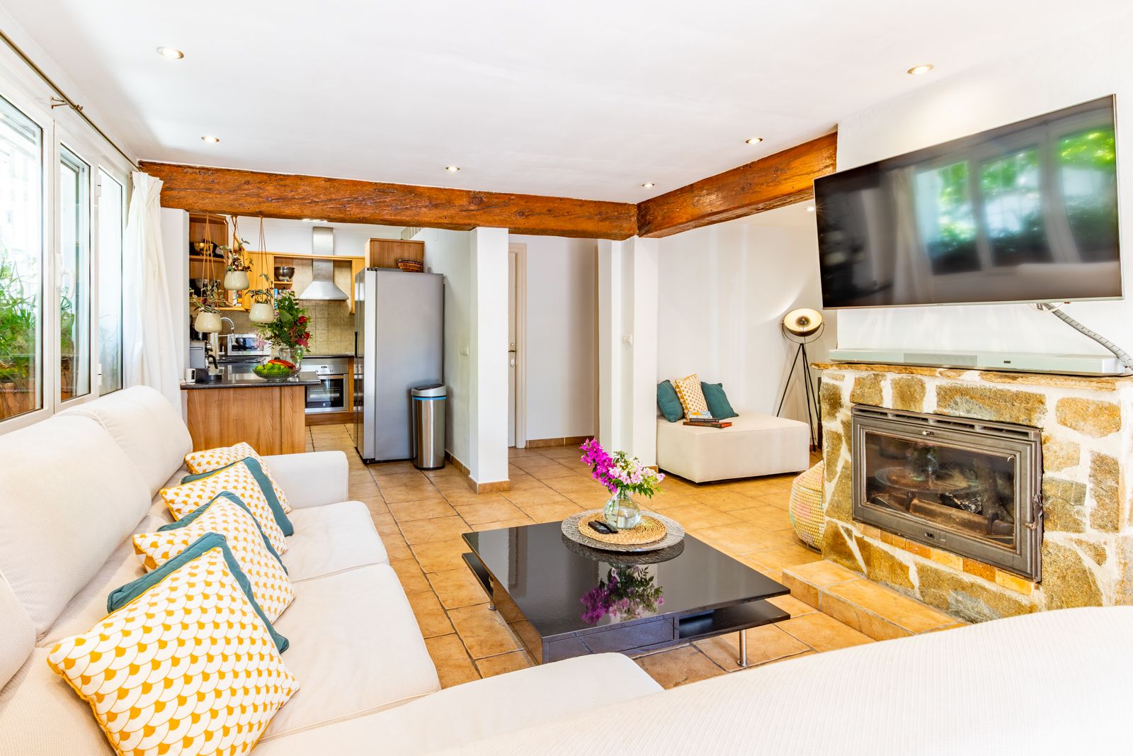 Bright living room featuring exposed wooden beams, a natural stone fireplace, and ceramic flooring, seamlessly connected to the open-plan kitchen.