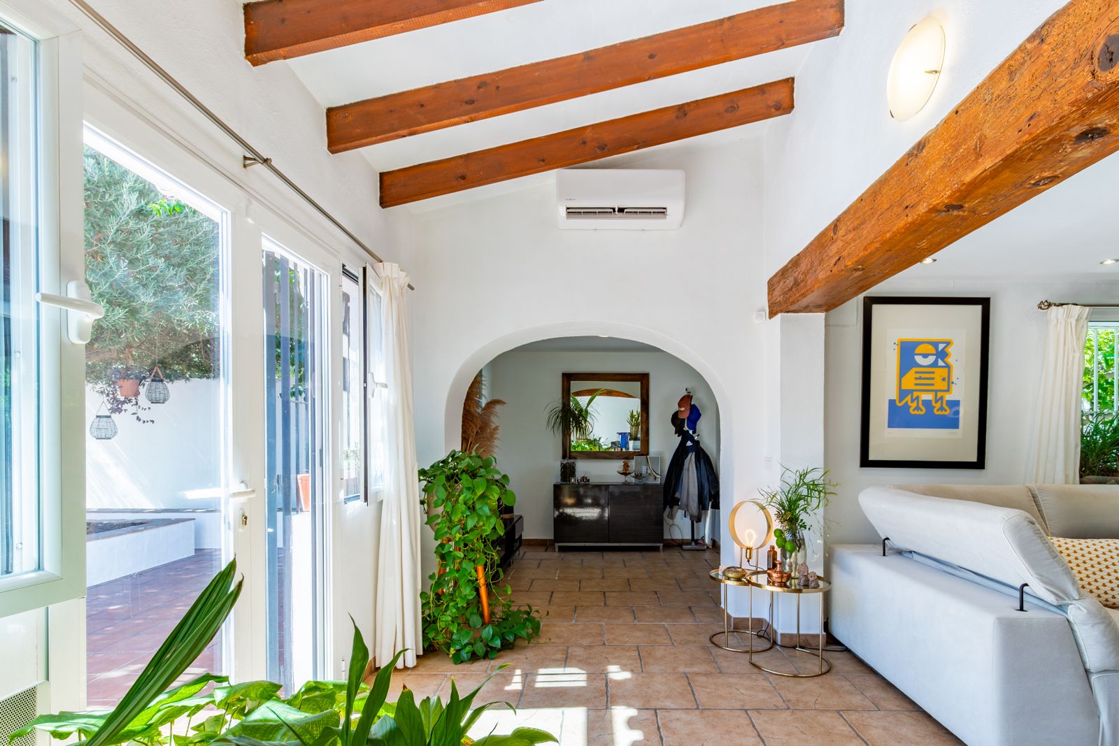 Bright living space featuring exposed wooden beams, terracotta flooring, and large glass doors. An arched entryway connects to the hall under a sloped ceiling with air conditioning.
