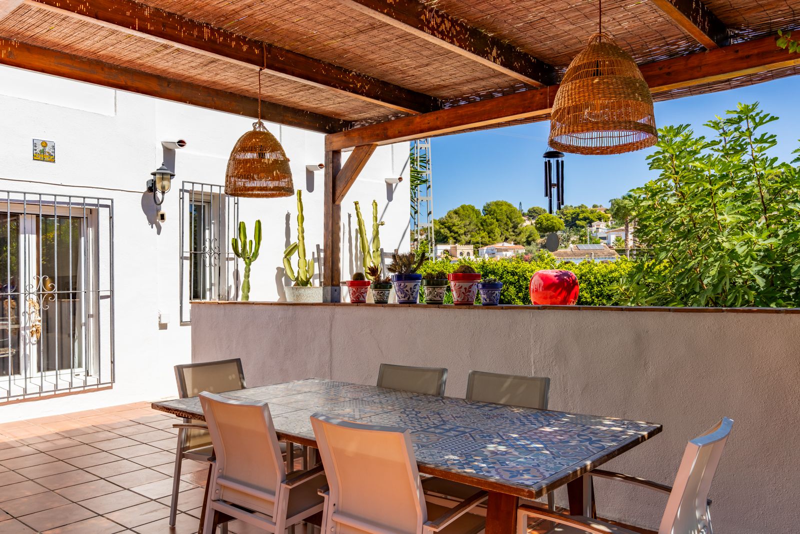 Covered terrace featuring a wooden pergola with reed roofing, a tiled dining table, and wicker pendant lights overlooking the Mediterranean landscape.