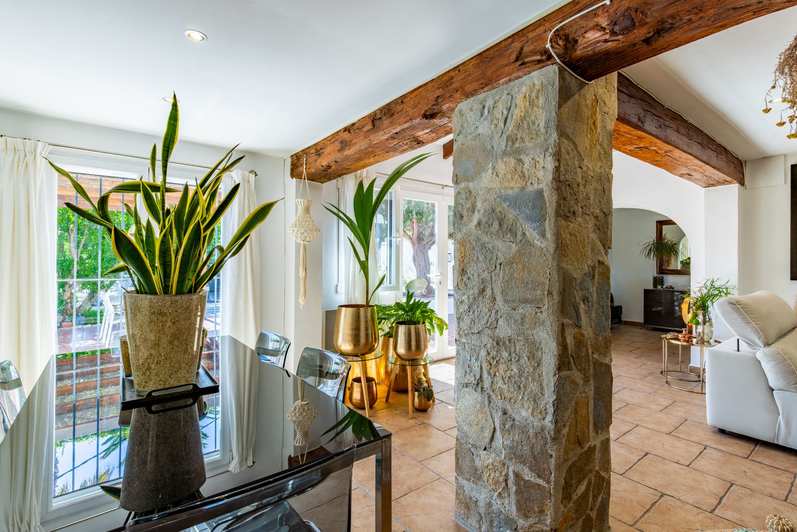 Dining space featuring exposed wooden beams and a natural stone pillar. Includes terracotta flooring, a glass table, and large windows for natural light.