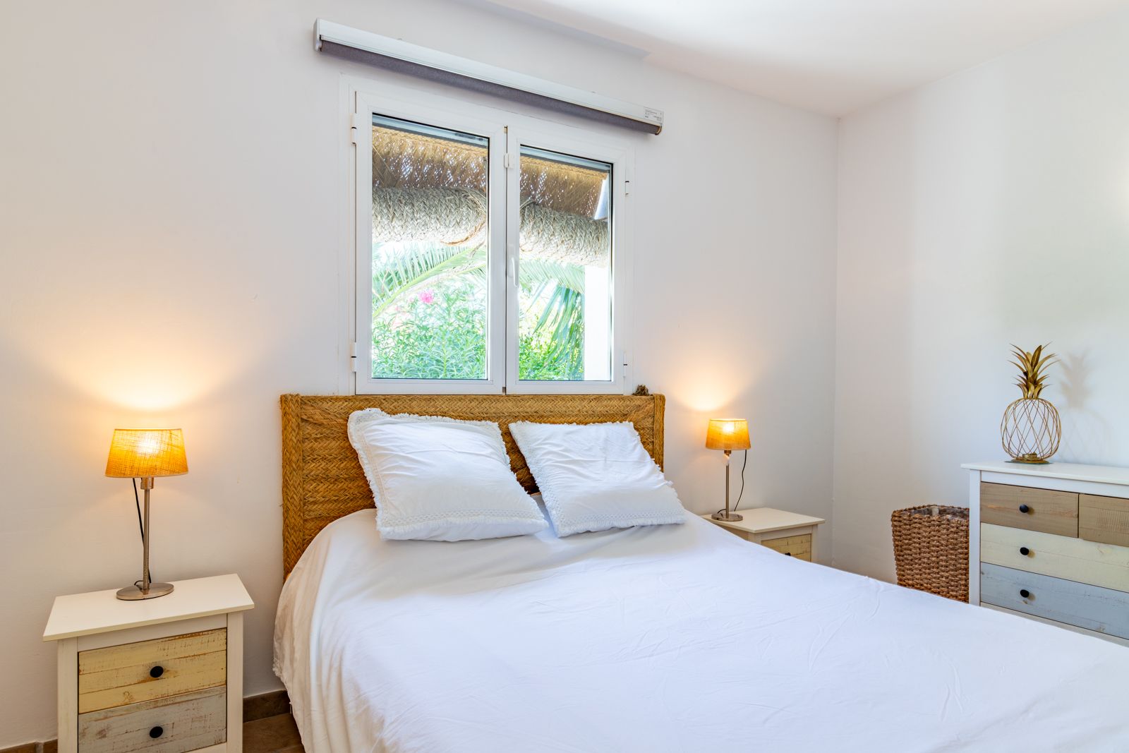Double bedroom featuring a wicker headboard, rustic wooden nightstands, and a window with garden views. A bright space with Mediterranean charm.