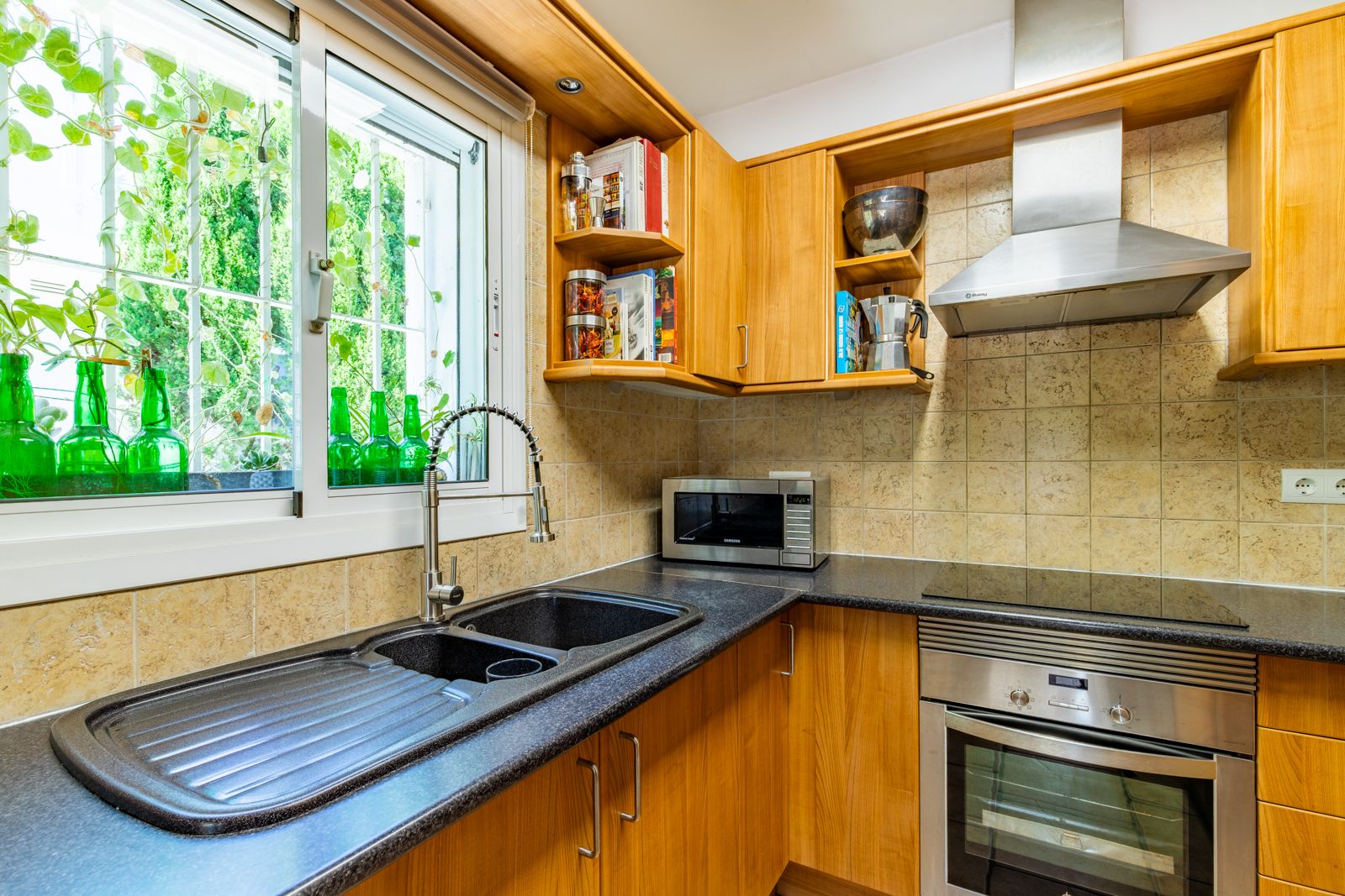 Fully equipped kitchen with wooden cabinetry, dark granite countertops, and a double sink. Features stainless steel appliances and natural light from the garden window.