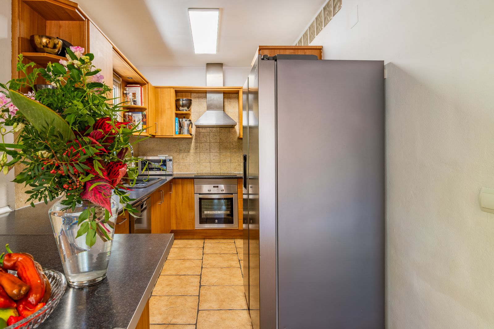 Functional kitchen featuring warm wood cabinetry, dark countertops, and stainless steel appliances. Includes a large refrigerator and terracotta-toned tiled flooring.