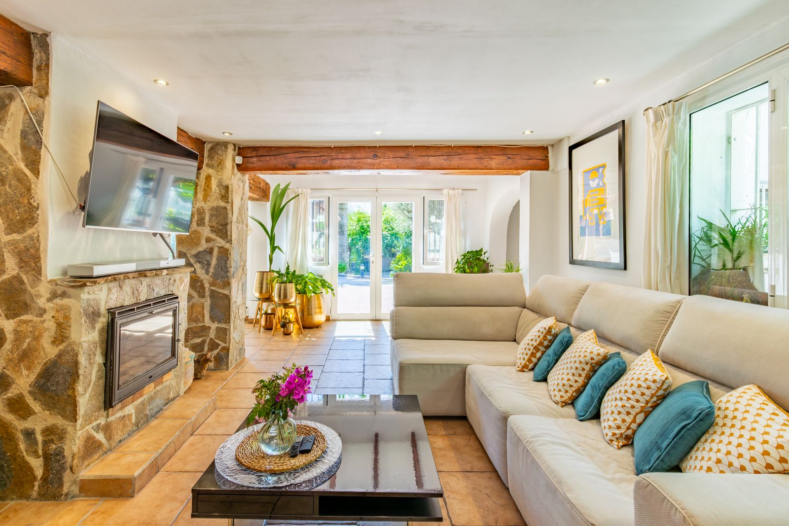 Living room with a stone-clad fireplace, exposed wooden beams, and terracotta tiling. Large glass doors integrate the interior with the garden.